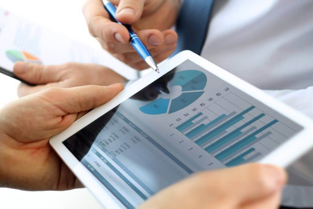 Two people review a tablet displaying a blue pie chart and bar graphs while one holds a pen.